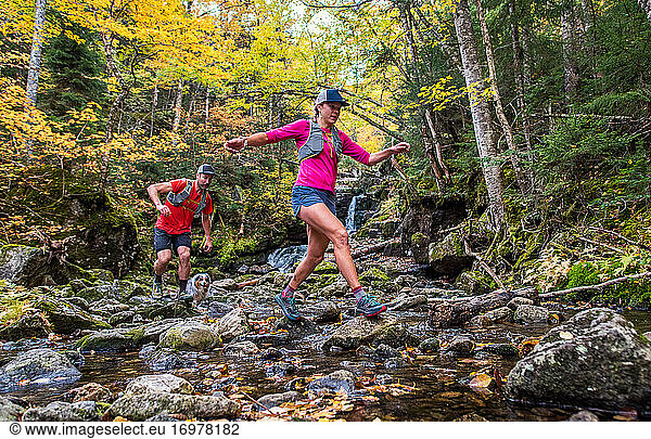 Man and woman trail runners rock-hopping in a river with fall foliage