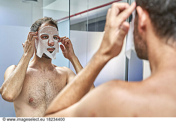 Man applying sheet mask on face in front of mirror