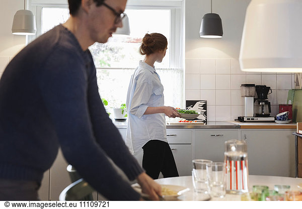 Man arranging dining table while woman preparing food at kitchen