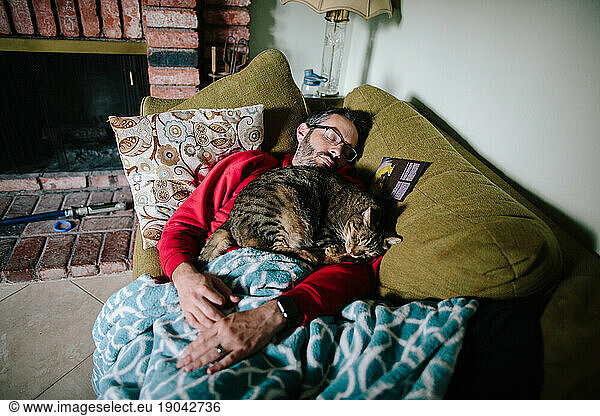 Man asleep on a couch with a brown tabby cat asleep on him