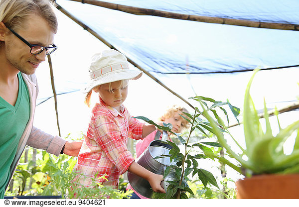 Man assisting girl watering plants with watering can in greenhouse  boy in background