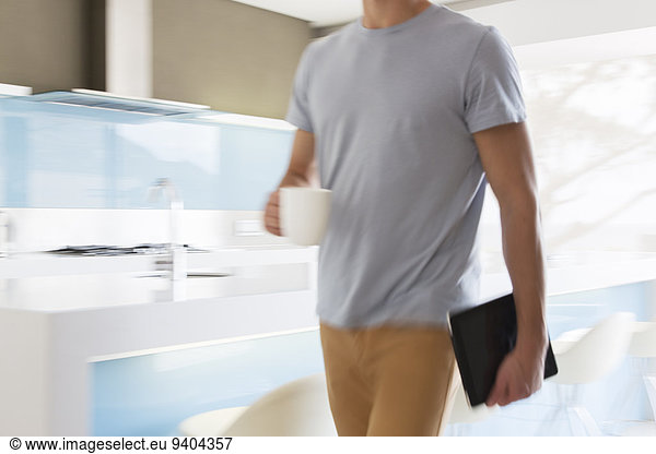 Man carrying coffee cup and digital tablet walking through modern kitchen