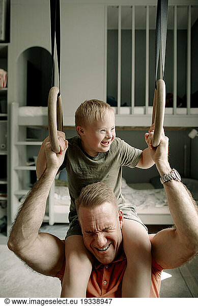 Man carrying son with down syndrome on shoulders while holding gymnastic rings in bedroom