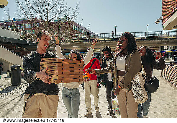 Man carrying stack of pizza boxes while walking with friends at street on sunny day