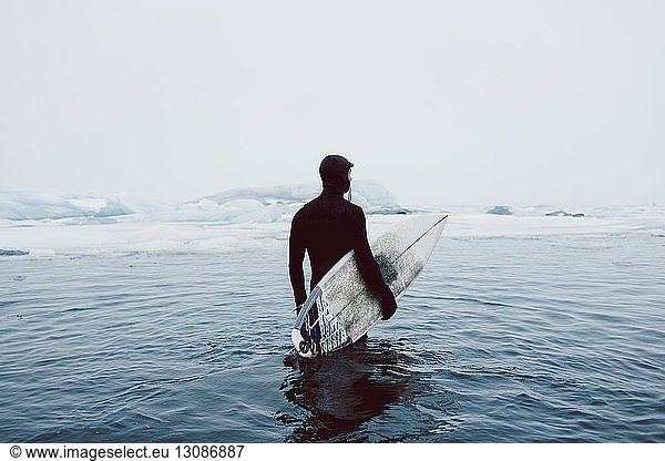 Man carrying surfboard while standing in icy sea against clear sky during winter
