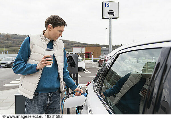 Man charging car holding disposable cup at station