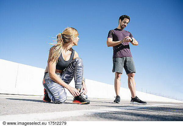 man checking time while woman tying shoelace on road against clear blue sky in city