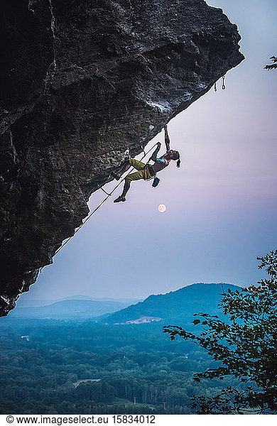 Man clipping quickdraw on overhanging sport climbing route with moon.