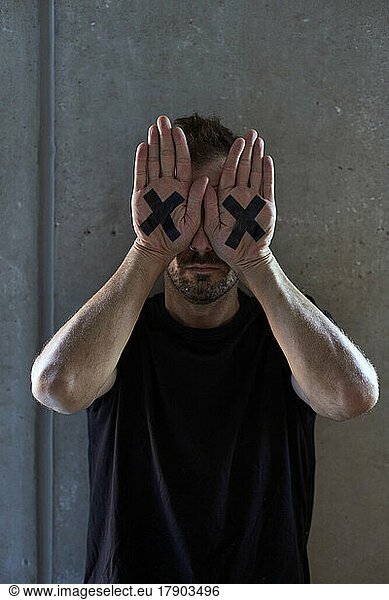 Man covering eyes with hands having cross marking in front of wall