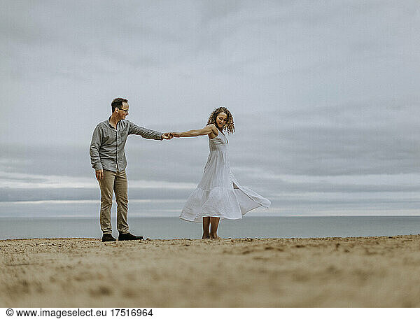 Man dances with woman in white dress on sandy beach next to water