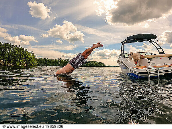 Man diving off boat into a lake on summer day as dog watches.