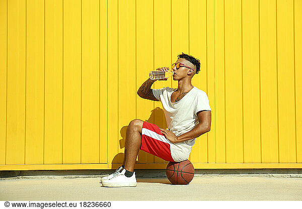 Man drinking water from bottle sitting on basketball in front of yellow wall