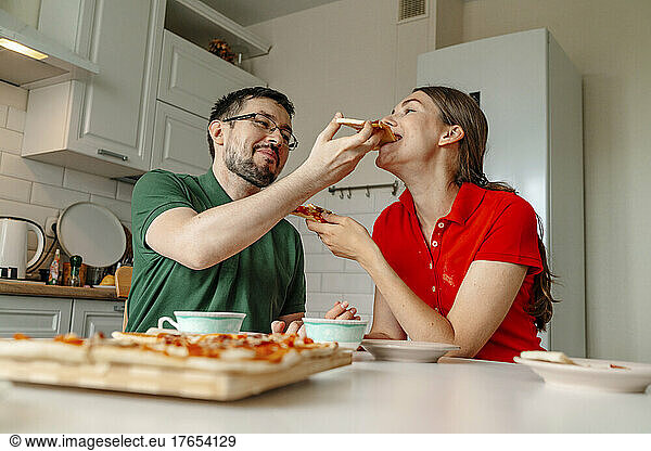 Man feeding pizza to woman sitting at table in kitchen