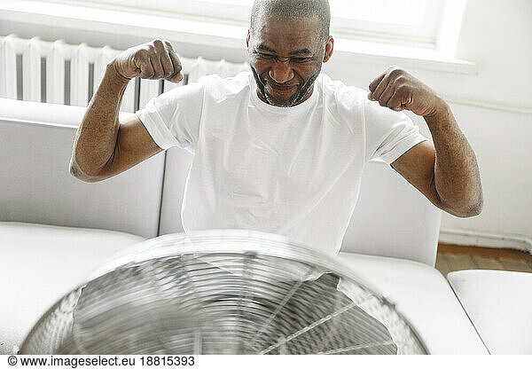 Man flexing muscles in front of electric fan at home