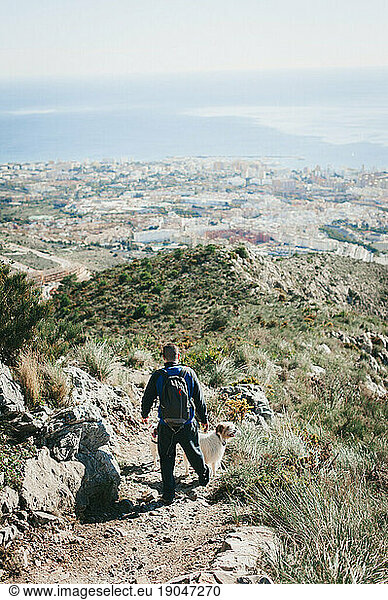 Man hiking down a hill with dog overlooking city and ocean in Malaga