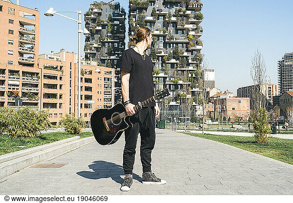 man holding a guitar is standing in front of two green skyscreapers