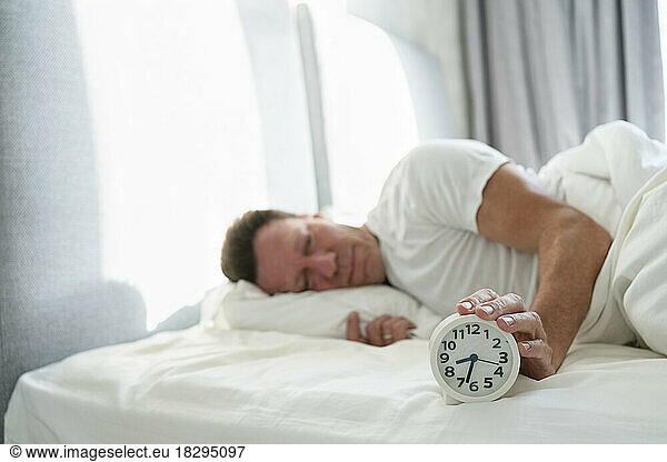 Man holding alarm clock lying on bed in bedroom
