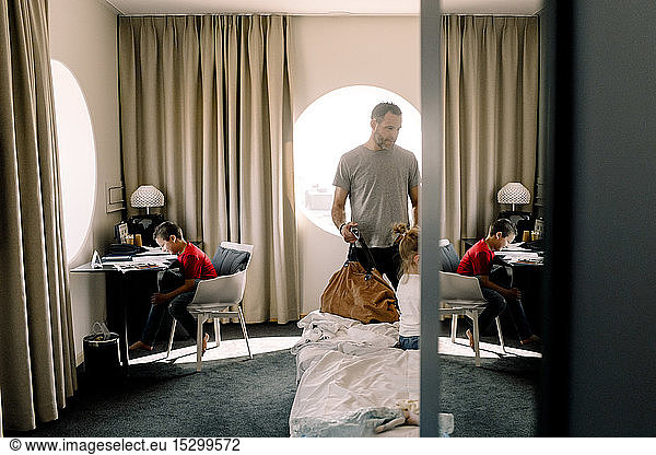 Man holding bag by bed while boy sitting at table in hotel