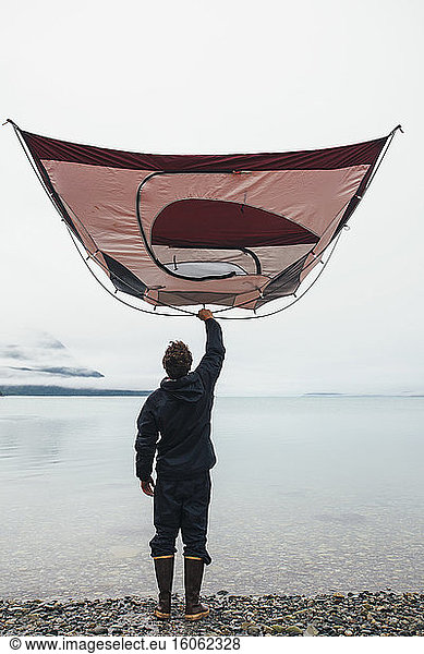Man holding camping tent over head standing on rocky beachan inlet on the Alaska coastline.