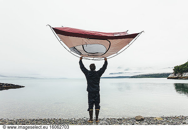 Man holding camping tent over head standing on rocky beachan inlet on the Alaska coastline.
