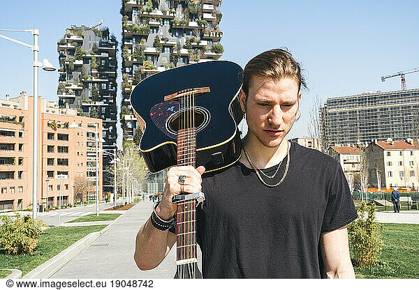 man holding guitar in his shoulder in front of two green skyscreapers