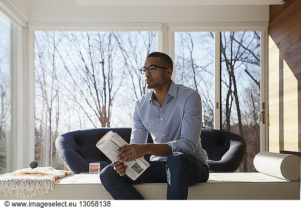 Man holding newspaper looking away while sitting on sofa at home