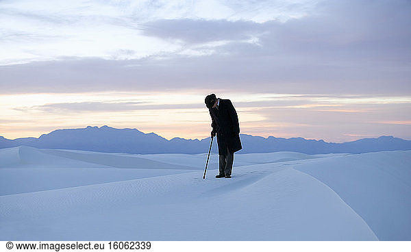 Man in a black coat and suit a bowler hat and umbrella in a white desert wilderness of white sand.