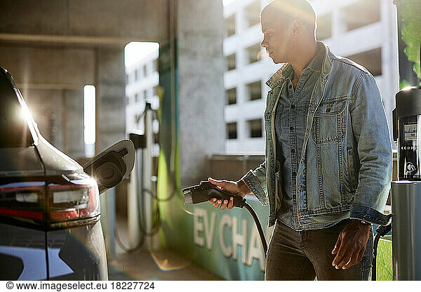 Man in denim jacket holding electric plug by car at charging station
