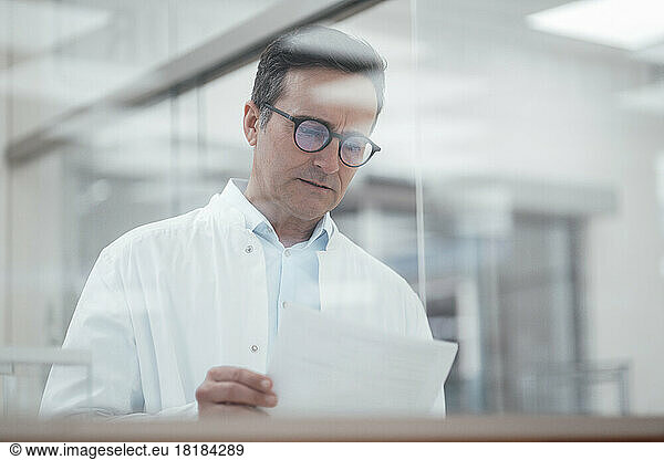 Man in lab coat analyzing medical reports in laboratory seen through glass