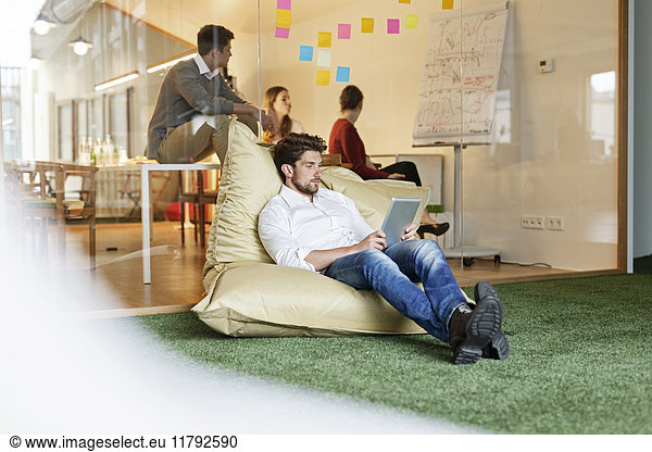 Man in office using tablet in bean bag with meeting in background
