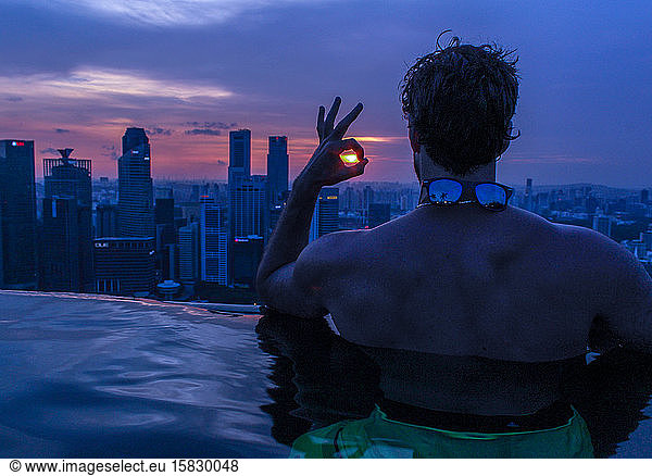 Man in rooftop infinity pool at sunset looking out at skyscrapers.