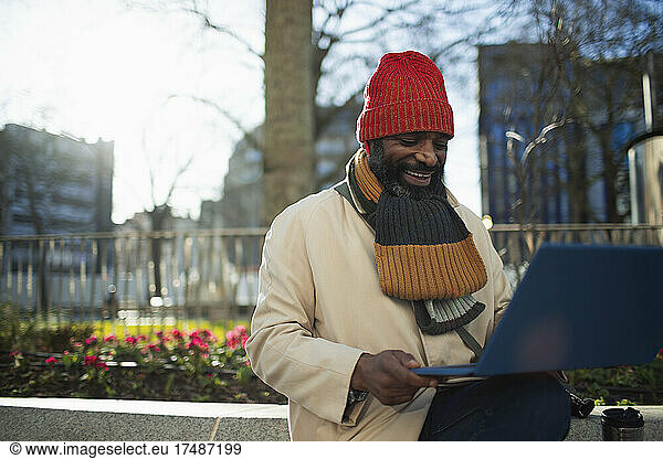 Man in stocking cap and scarf using laptop in sunny city park