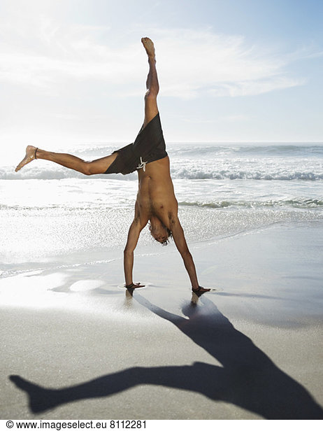 Man in swim trunks doing handstand on beach