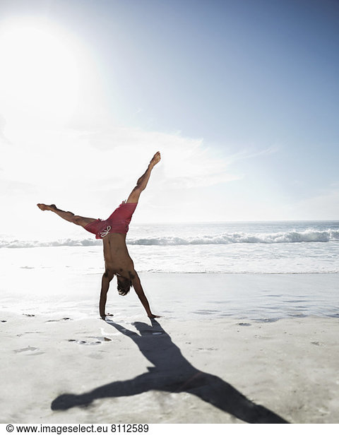 Man in swim trunks doing handstand on beach