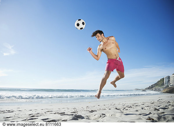 Man in swim trunks heading soccer ball on beach
