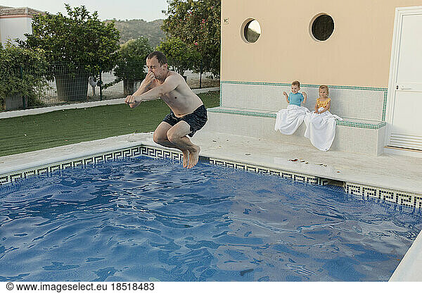 Man jumping in swimming pool with son and daughter sitting in background
