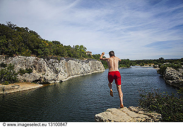 Man jumping off cliff into river against sky