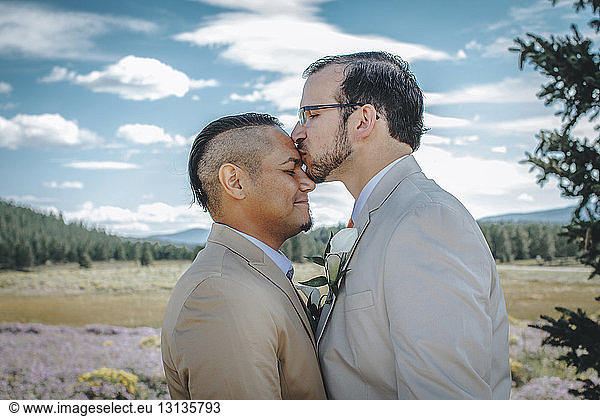 Man kissing boyfriend on forehead while standing against cloudy sky
