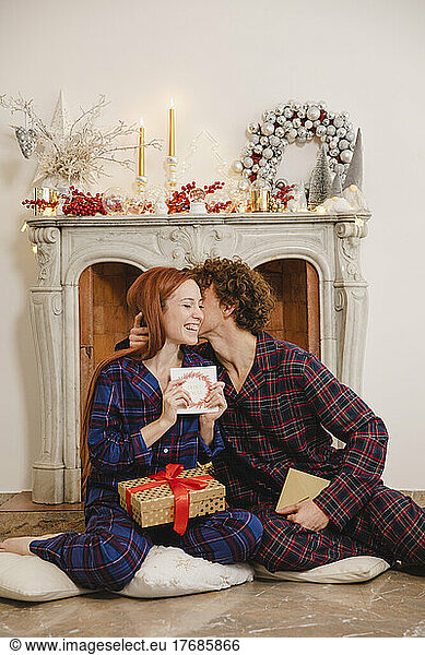 Man kissing girlfriend holding greeting card sitting with gift in front of fireplace at home