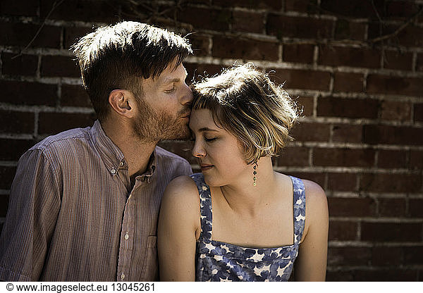 Man kissing girlfriend on forehead while standing against brick wall