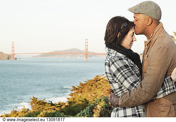Man kissing girlfriend's forehead while embracing against bridge