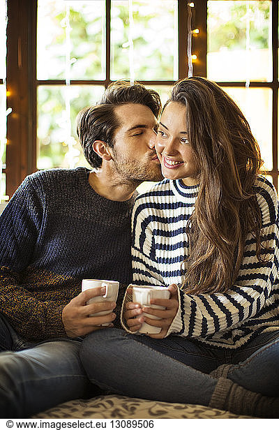 Man kissing girlfriend while having coffee on alcove window seat at home
