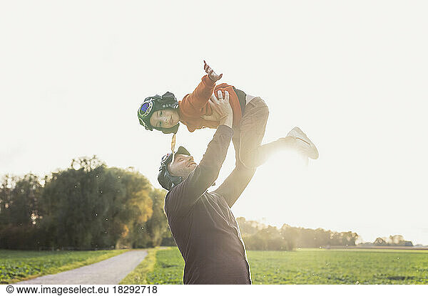 Man lifting son wearing flying goggles in autumn