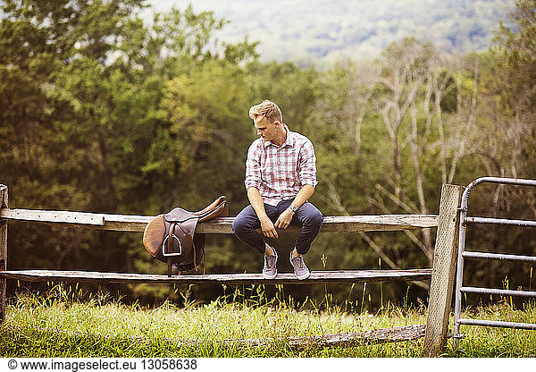 Man looking at saddle while sitting on railing in farm