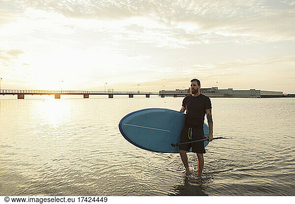 Man looking away carrying paddleboard in sea during sunset