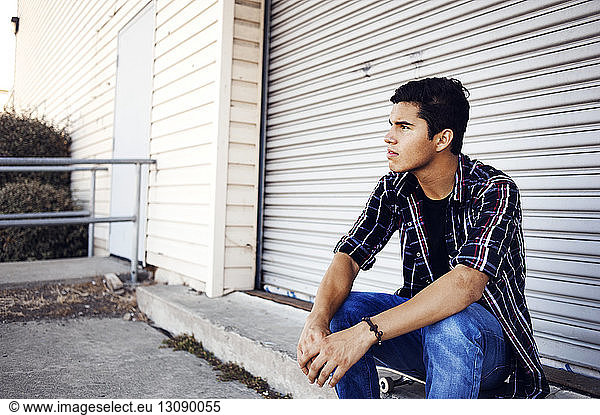 Man looking away while sitting on skateboard against shutter