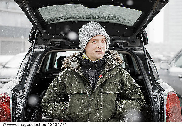 Man looking away while standing by car trunk during snowfall
