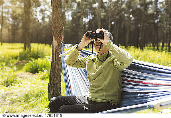 Man looking through binoculars sitting in hammock on sunny day