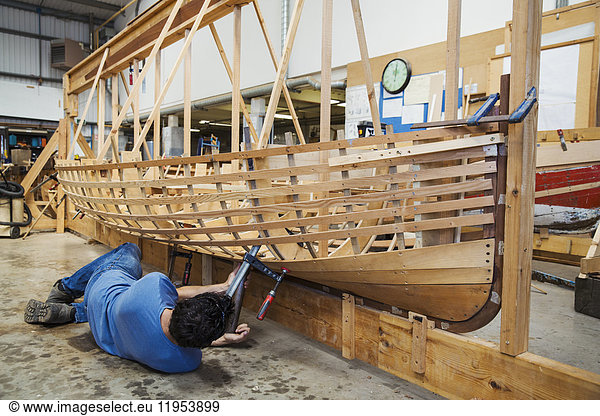 Man lying on floor in a boat-builder's workshop  working on a wooden boat hull.