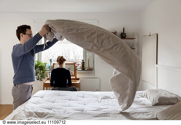 Man making bed while woman working at table in bedroom